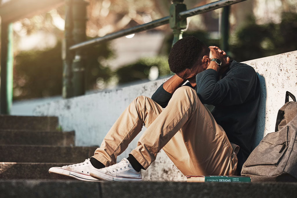 Distraught man sitting on a sidewalk with his head in his hands showing signs of needing mental health treatment